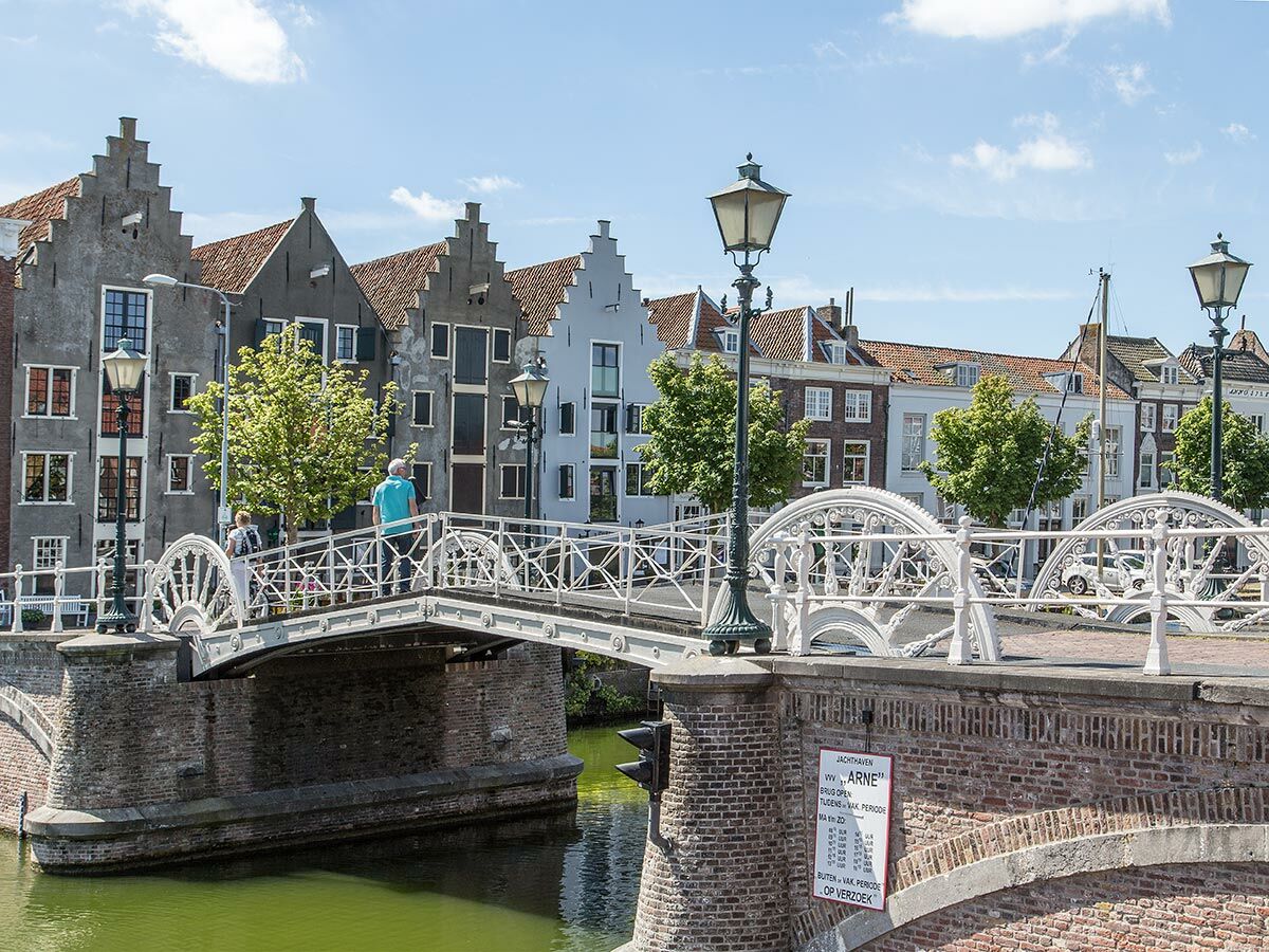 Blick auf eine historische Grachtenstadt in den Niederlanden mit weißer Brücke, Giebelhäusern und Fußgängern bei Sonnenschein.