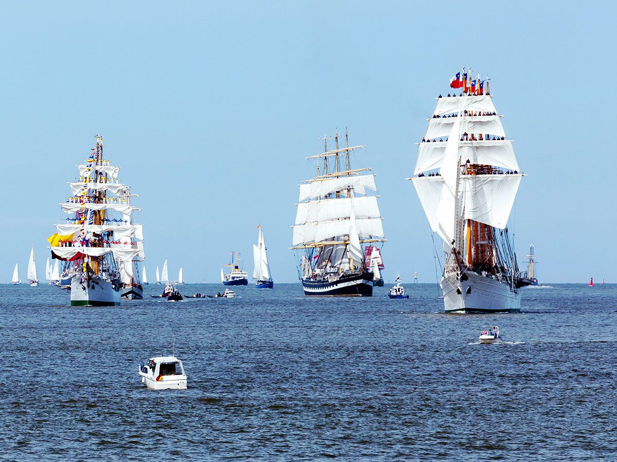 Drei majestätische Großsegler mit weißen Segeln und vielen Zuschauern fahren auf offenem Meer, umgeben von kleineren Booten.