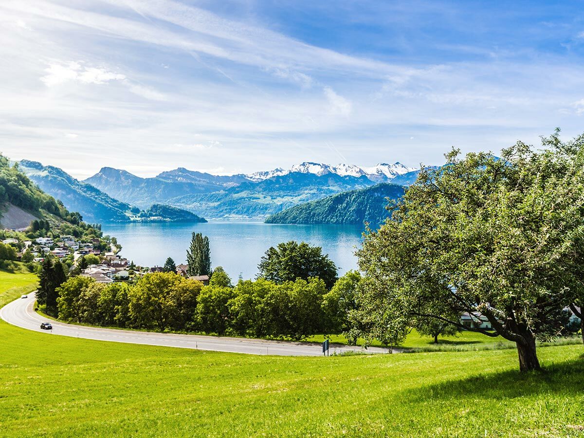 Panoramabild einer malerischen Bergsee-Landschaft mit schneebedeckten Gipfeln, grünem Uferdorf, See und kurviger Landstraße.