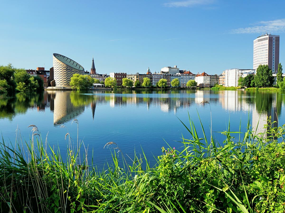 Kopenhagens Panorama: Blauer Planet, Kirche, Stadtgebäude am Seeufer unter blauem Himmel, mit Reflexionen und grünen Pflanzen.