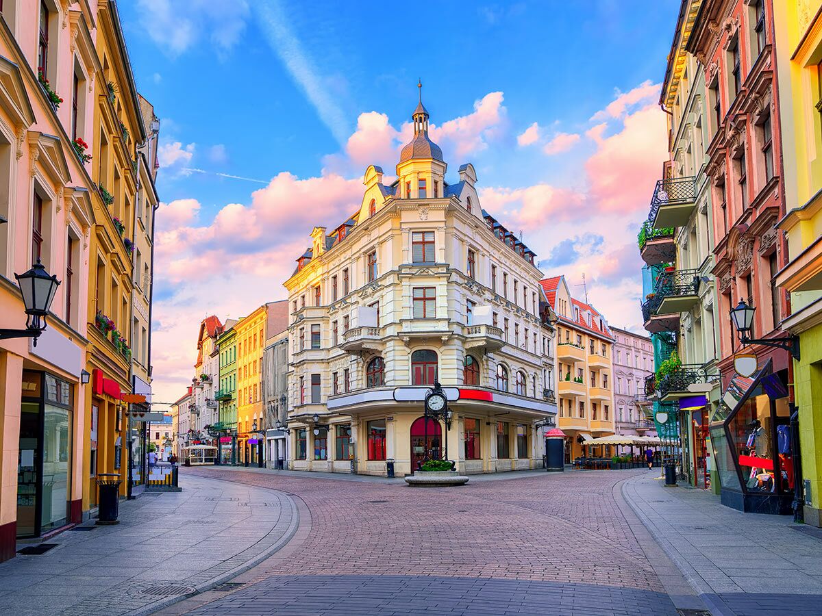 Eine malerische europäische Altstadtstraße mit bunten historischen Gebäuden, Geschäften und einem markanten Eckhaus unter rosa Wolken.