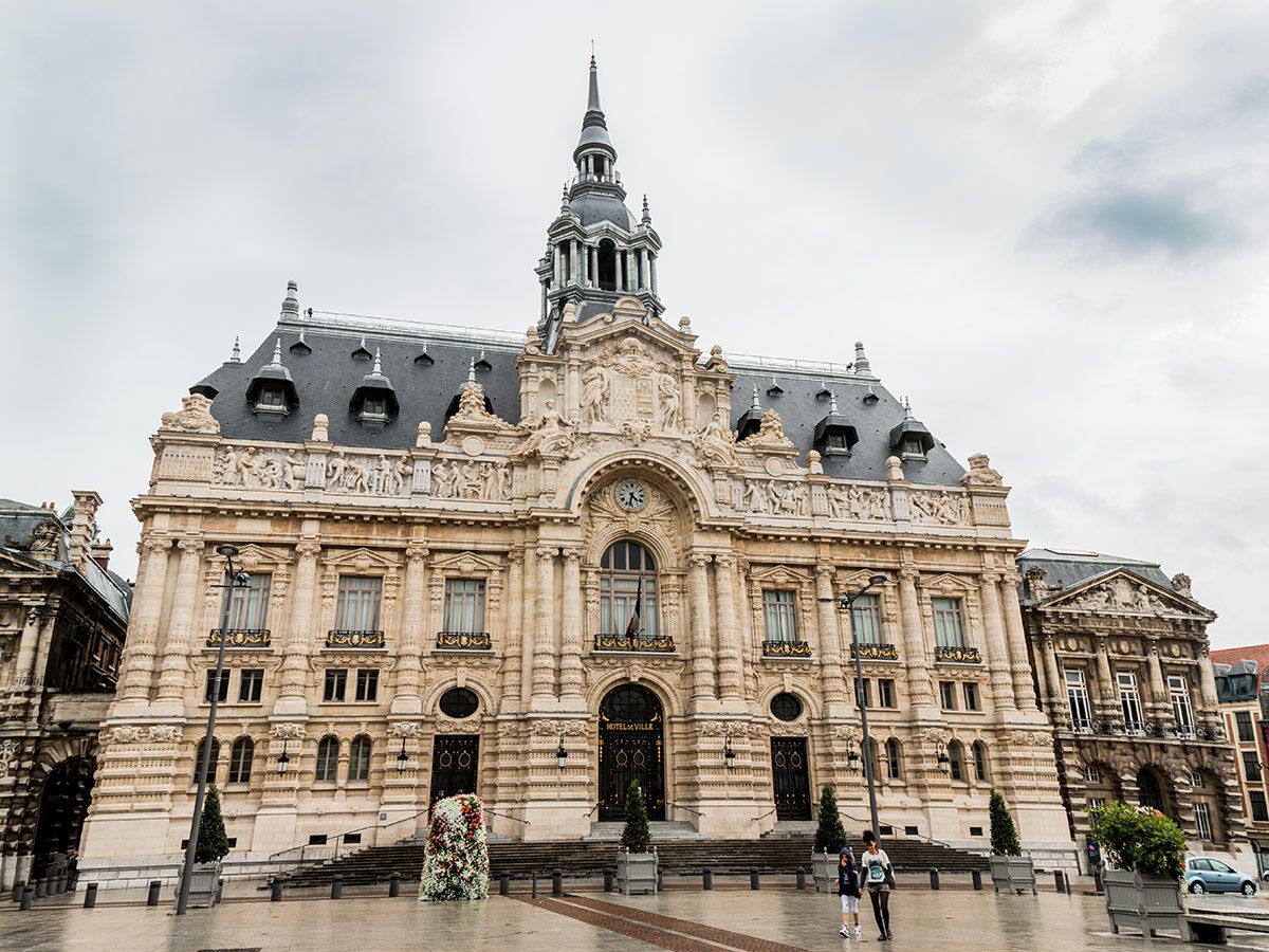 Hôtel de Ville Calais mit aufwendiger Fassade, Turm, Uhr und Skulpturen unter bewölktem Himmel. Zwei Personen auf dem Platz.