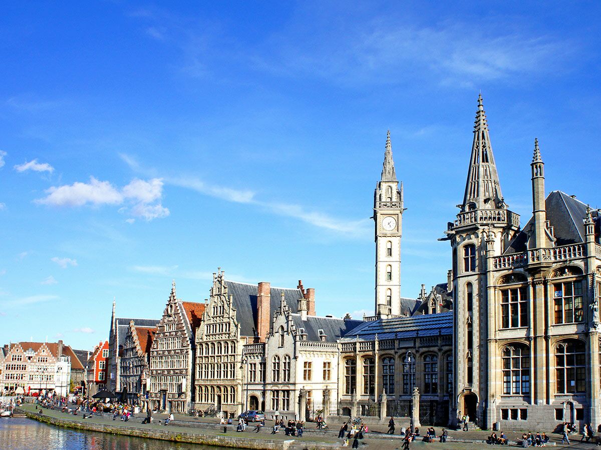 Panoramablick auf das historische Gent mit Belfried, alten Giebelhäusern und Menschen entlang des Leie-Kanals unter blauem Himmel.