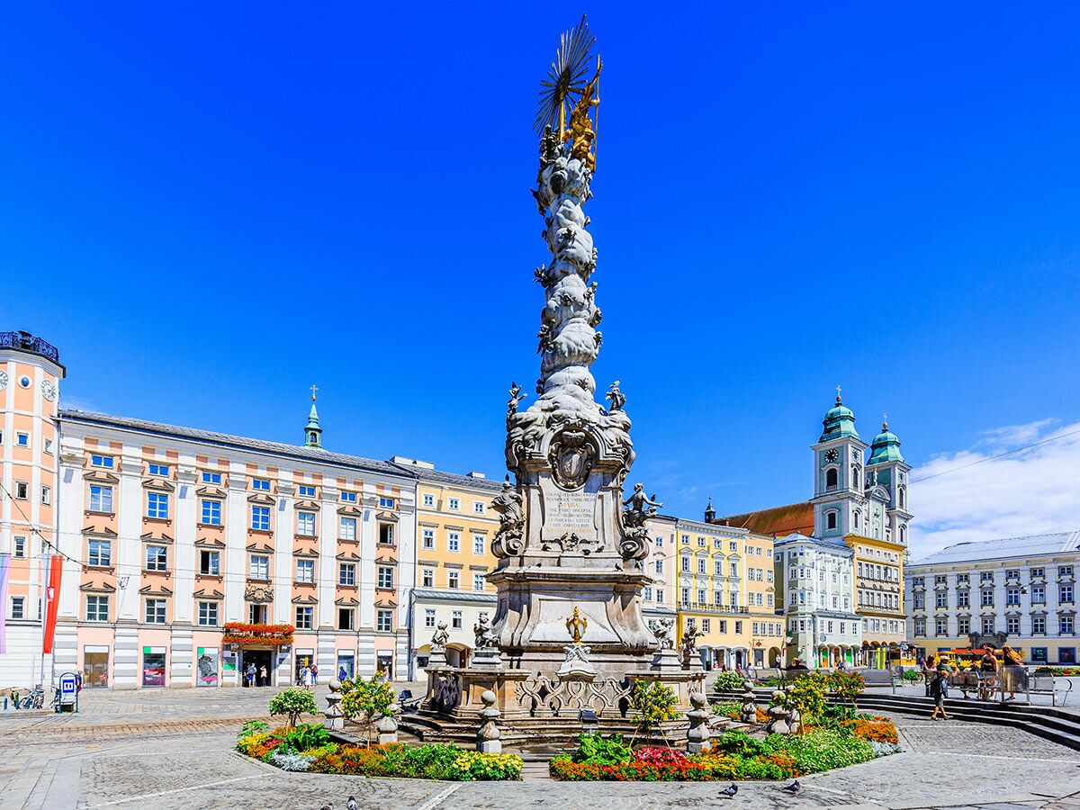 Blick auf den Linzer Hauptplatz mit der barocken Dreifaltigkeitssäule und dem Alten Dom unter blauem Himmel an einem sonnigen Tag.