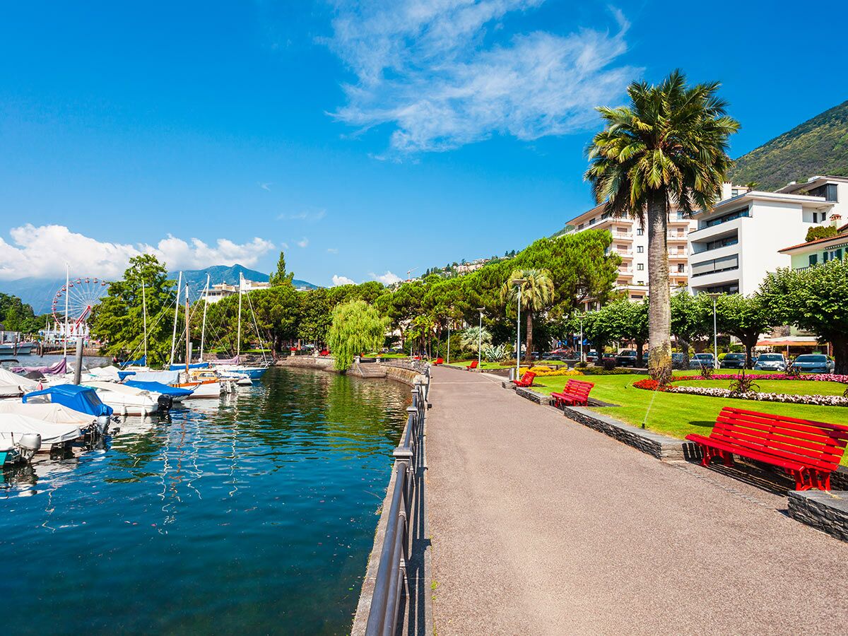 Blick auf eine sonnige Seeuferpromenade mit festgemachten Booten, roten Bänken, grünen Bäumen und Bergen im Hintergrund. Ein Riesenrad ist sichtbar.