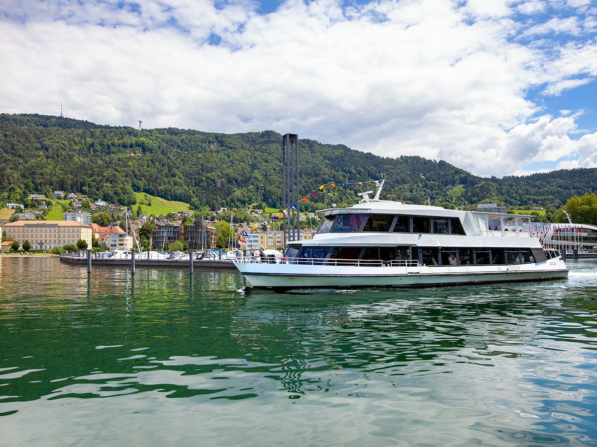 Weißes Passagierschiff fährt auf ruhigem See, umgeben von grünen Bergen und einer Stadt mit Hafen unter bewölktem Himmel.