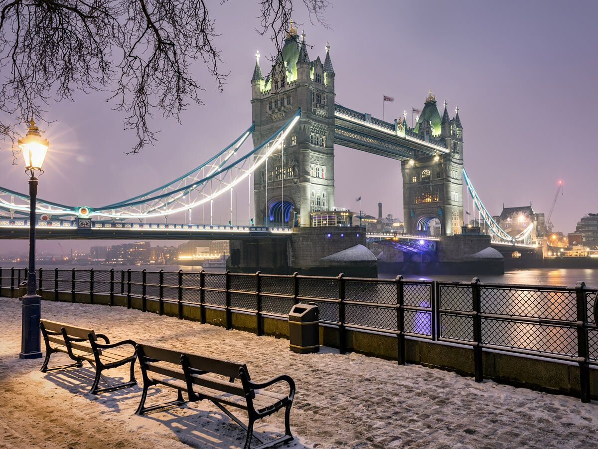 Die Tower Bridge in London bei Nacht im Winter, mit Schnee bedeckter Promenade, Bänken und Laterne am Flussufer.
