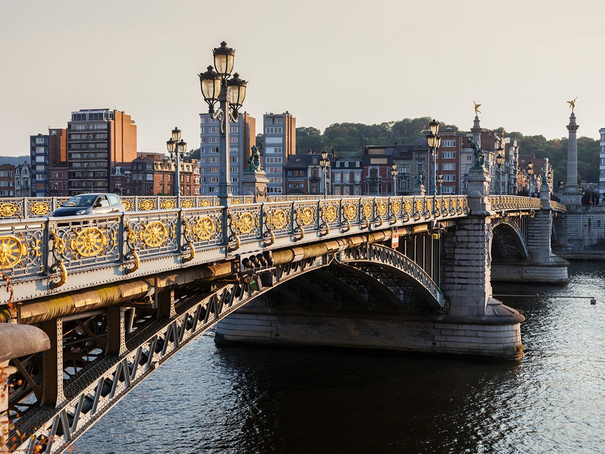 Goldverzierte Pont de Fragnée Brücke über die Maas in Lüttich mit einem Auto, Stadtgebäuden und Bäumen im warmen Abendlicht.