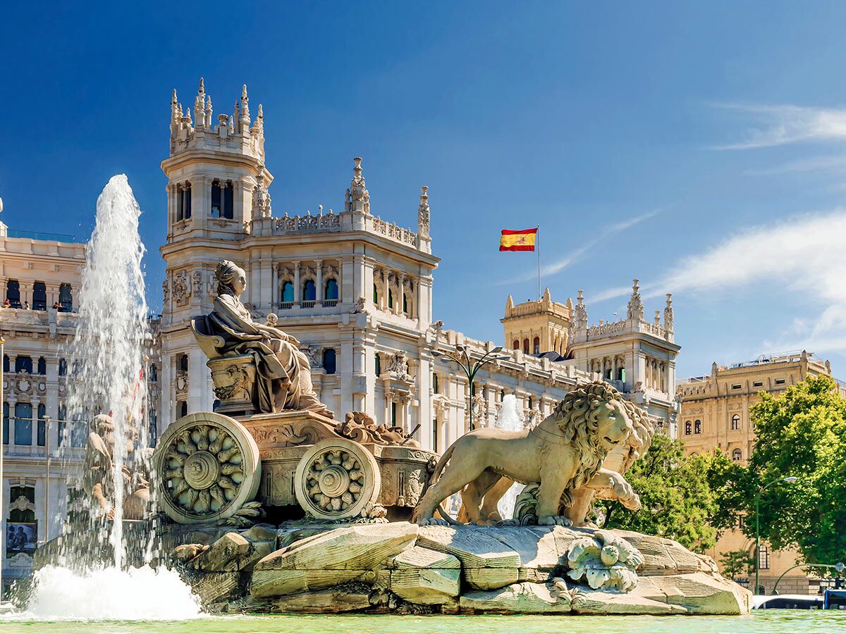 Der berühmte Cibeles Brunnen in Madrid mit der Göttin Kybele auf einem Löwenwagen vor dem beeindruckenden Palacio de Cibeles unter blauem Himmel.