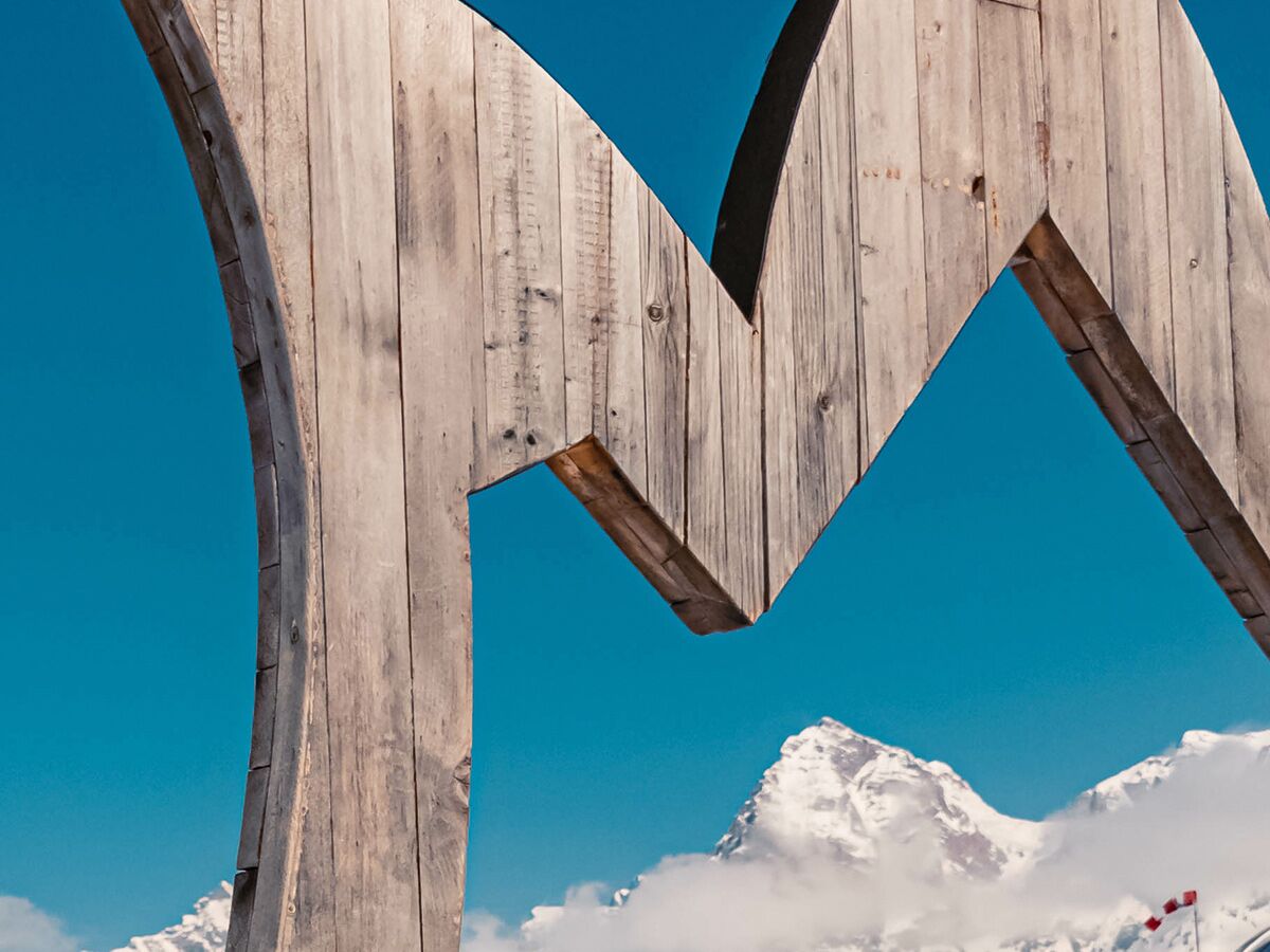 Große, M-förmige Holzkonstruktion vor blauem Himmel mit schneebedeckten Alpenbergen und Wolken im Hintergrund.