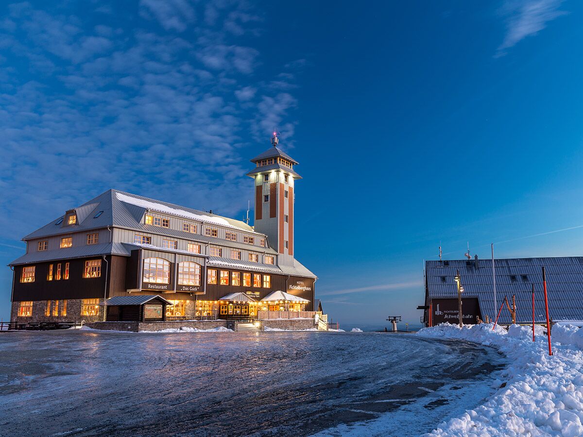 Das Fichtelberghaus und der Aussichtsturm im Winter bei Abenddämmerung mit Schnee und beleuchteten Fenstern.