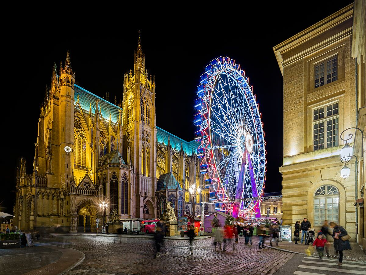 Die Metzer Kathedrale und ein leuchtendes Riesenrad bei Nacht mit bewegten Menschen auf dem festlichen Platz.