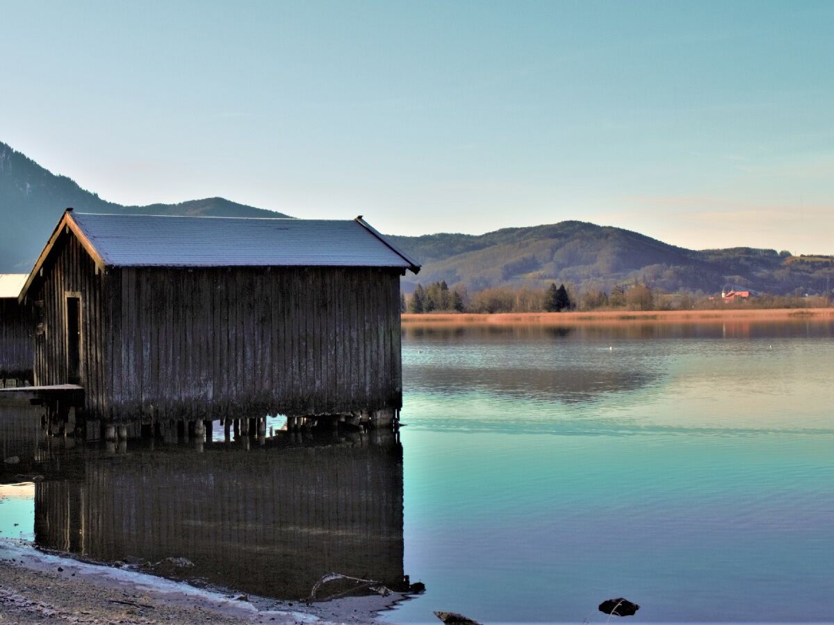 Altes Holz-Bootshaus mit Schneeschicht auf dem Dach am ruhigen Seeufer, umgeben von Bergen und Wäldern, mit Spiegelung im klaren Wasser.