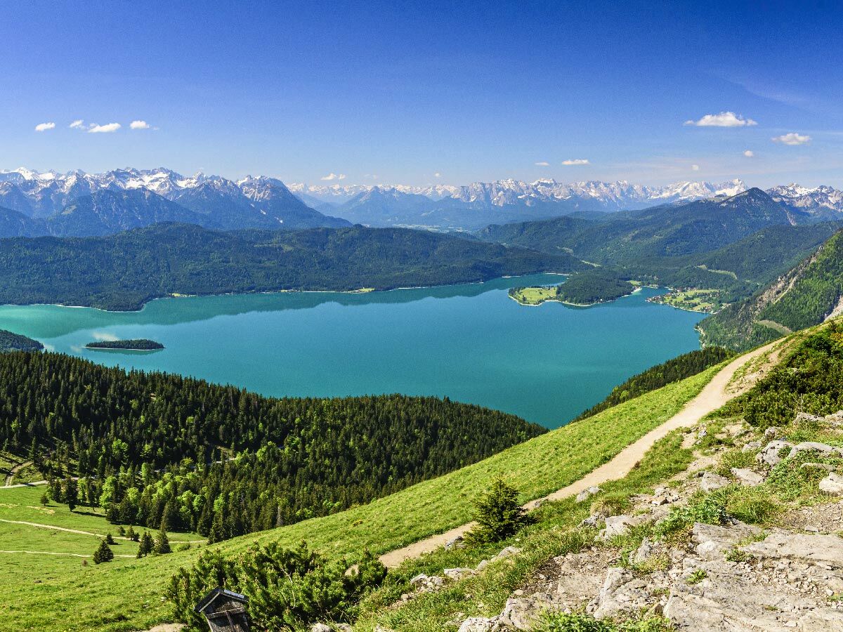 Atemberaubende Panoramaansicht eines türkisfarbenen Bergsees mit Insel, umgeben von dichten Wäldern und schneebedeckten Alpen, sowie einem Wanderweg.