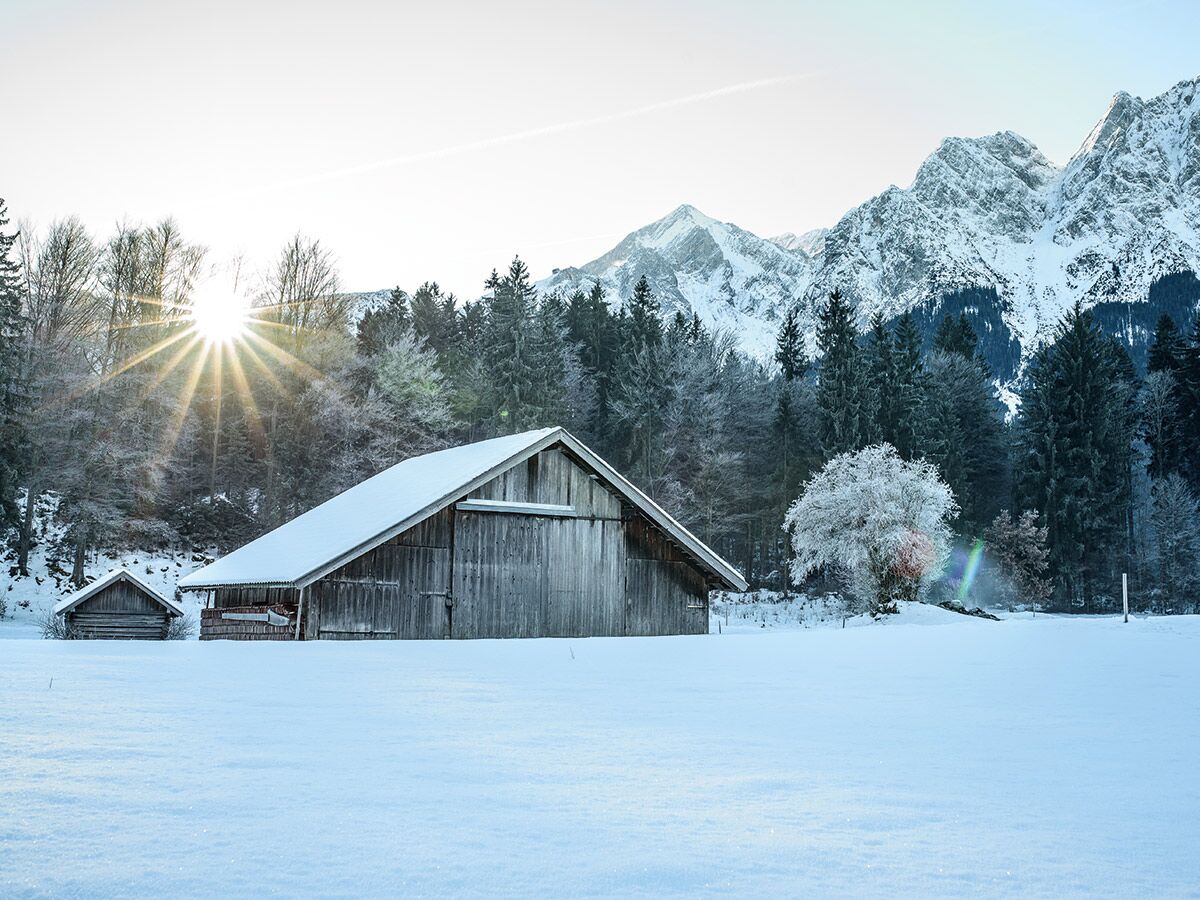 Holzscheune im tiefen Schnee vor verschneiten Bäumen und majestätischen Bergen mit strahlender Wintersonne.