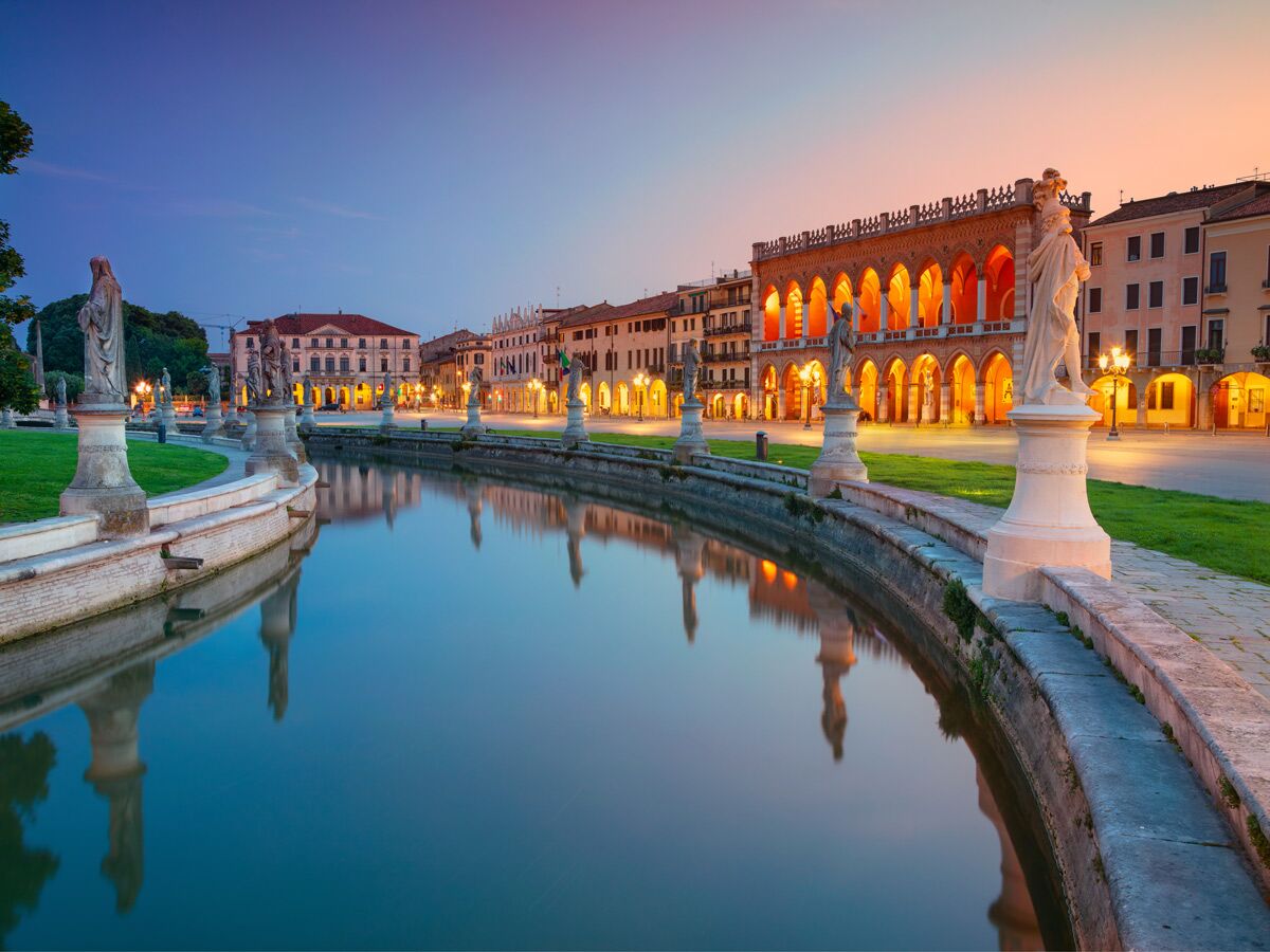 Prato della Valle in Padua bei Dämmerung, beleuchtete Gebäude und historische Statuen spiegeln sich im zentralen Kanal.