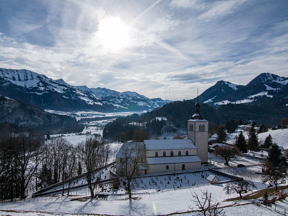 Schneebedeckte Kirche mit Friedhof vor majestätischer Alpenkulisse und sonnigem Himmel im Winter.