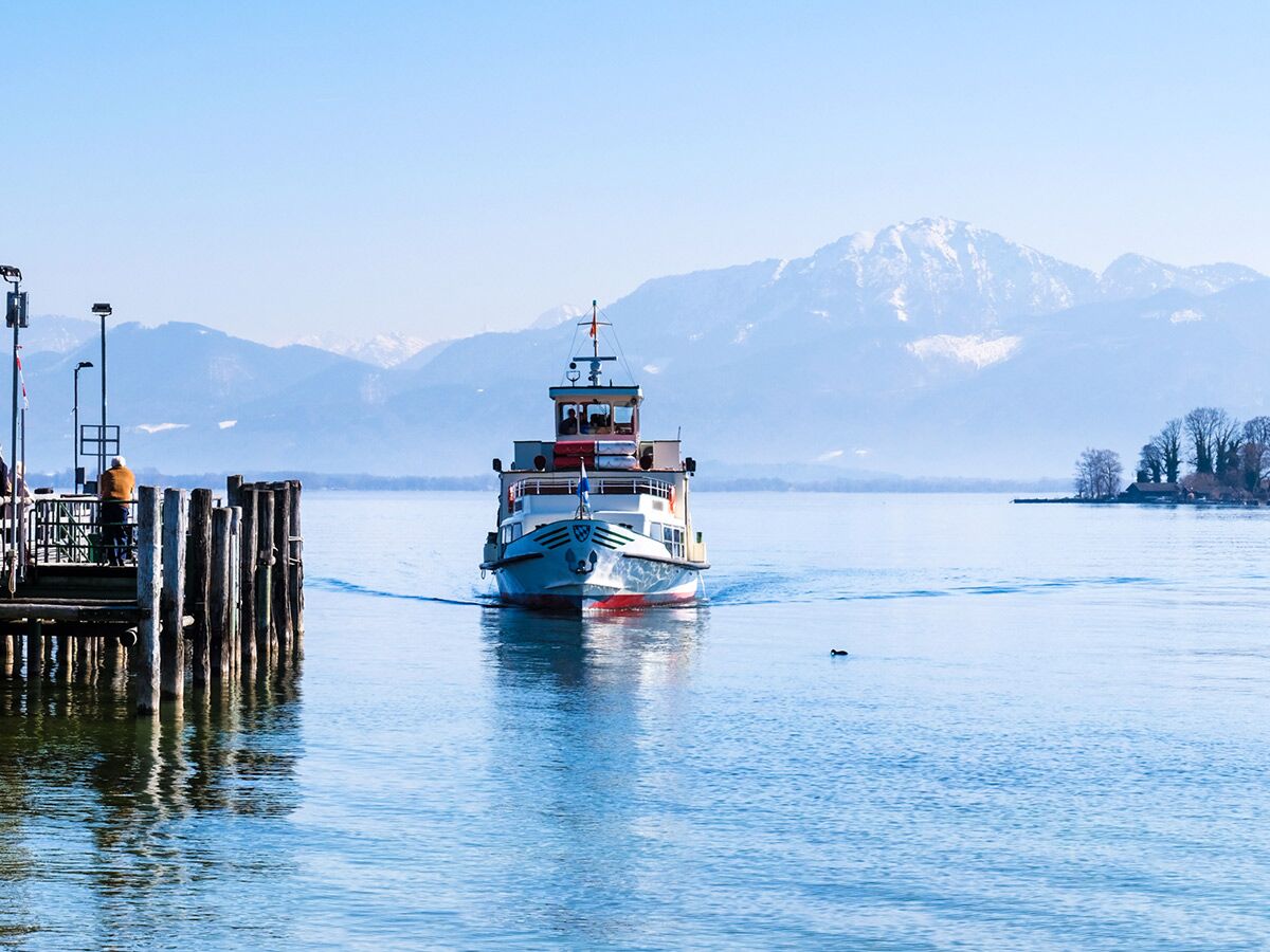 Fähre fährt auf blauem See mit schneebedeckten Bergen im Hintergrund und Holzpier im Vordergrund an einem klaren Wintertag.