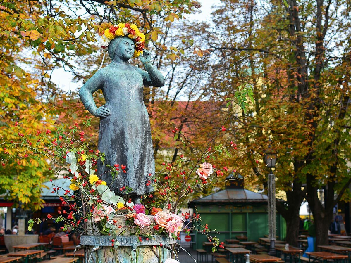 Bronze-Frauenstatue mit Blumenkranz und herbstlichen Blumen am Brunnen. Bunte Herbstbäume und Biergarten im Hintergrund.