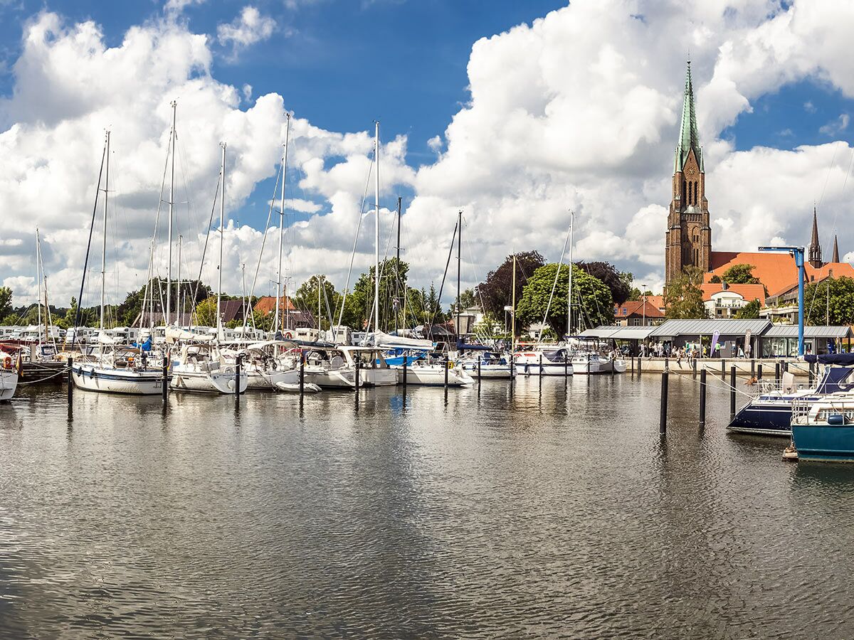 Malerische Marina mit Segelbooten, einer historischen Kirche und belebten Uferpromenade unter blauem Himmel mit Wolken.
