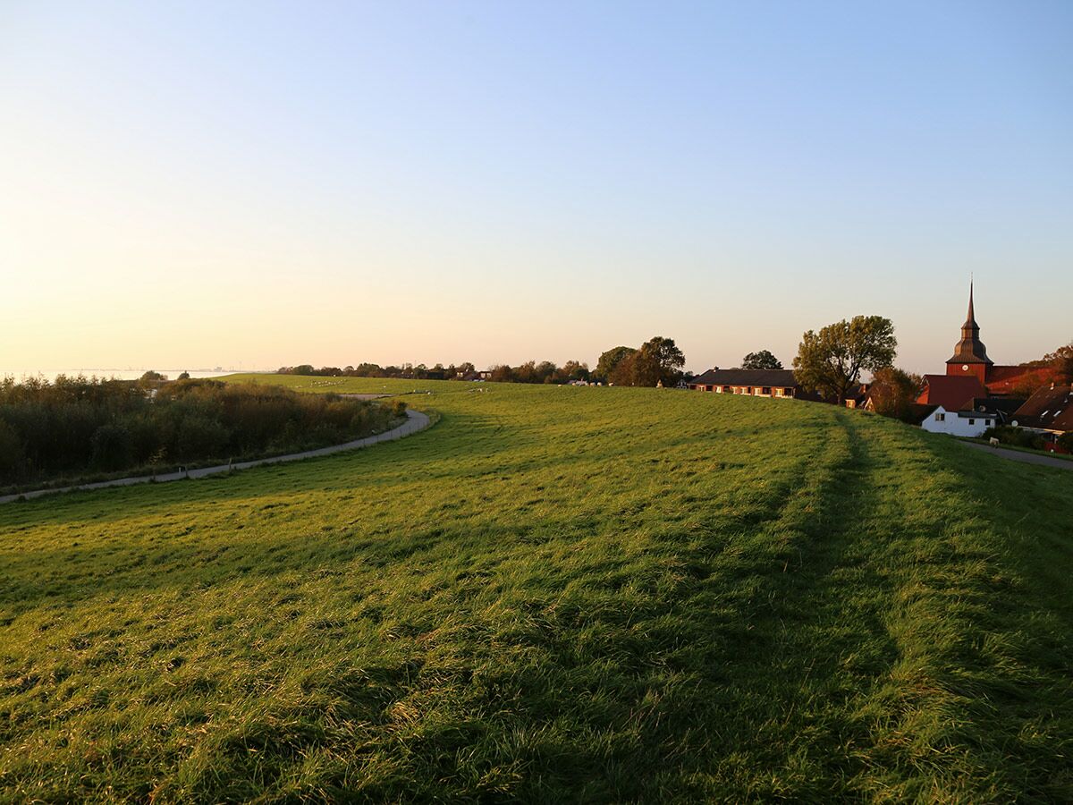 Grüne Deichlandschaft mit gewundenem Weg, Dorfkirche und Gewässer im warmen Abendlicht. Eine ruhige Küstenlandschaft.