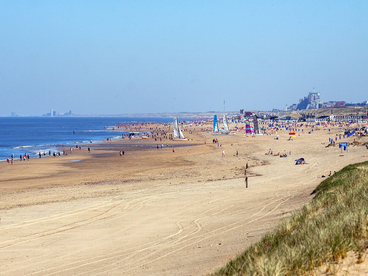 Panorama eines belebten Sandstrandes mit Menschen, bunten Katamaranen, ferner Stadtkulisse und Dünen unter blauem Himmel.