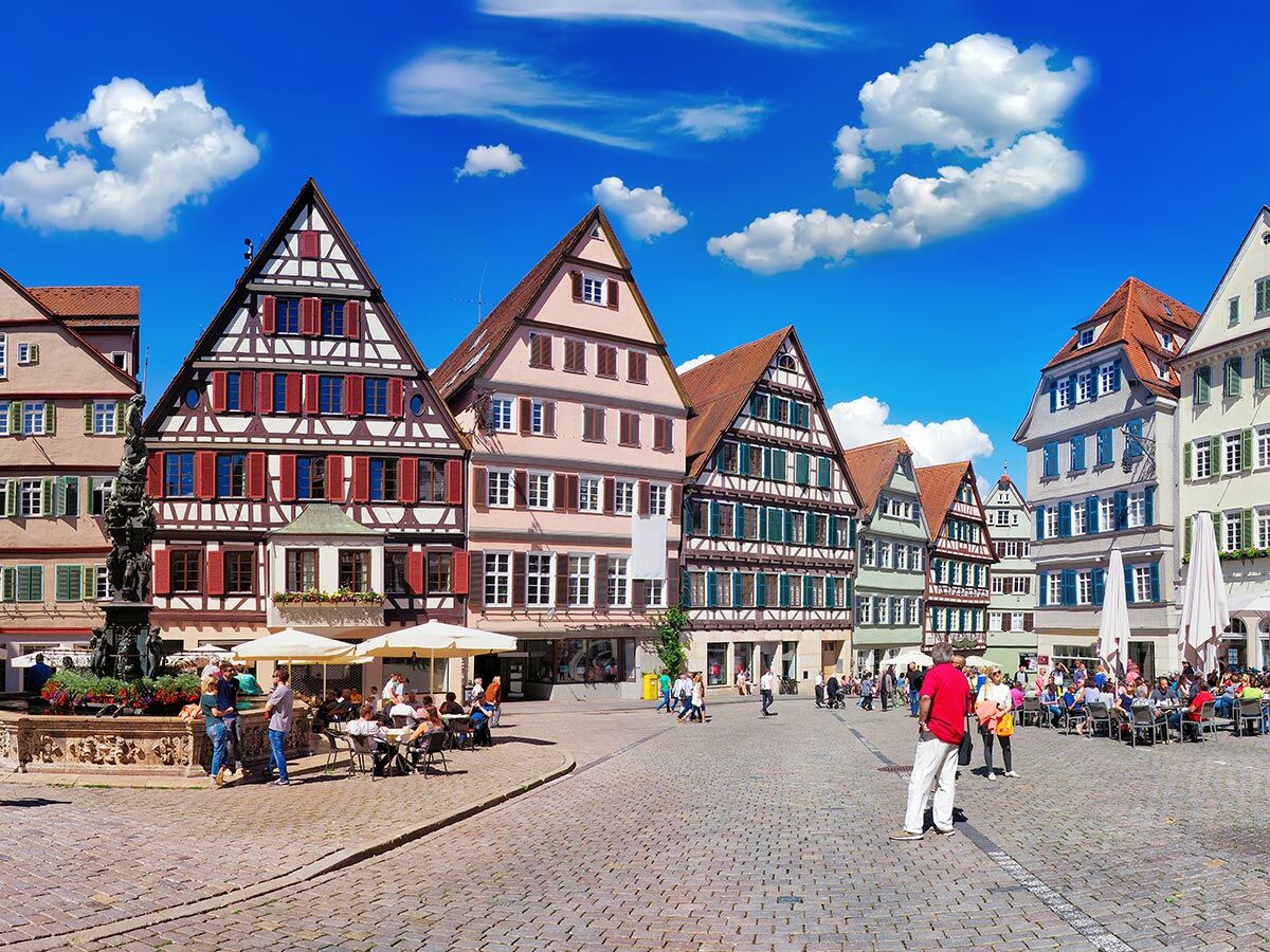 Blick auf einen belebten historischen Marktplatz mit traditionellen Fachwerkhäusern, Brunnen und Cafés unter blauem Himmel.
