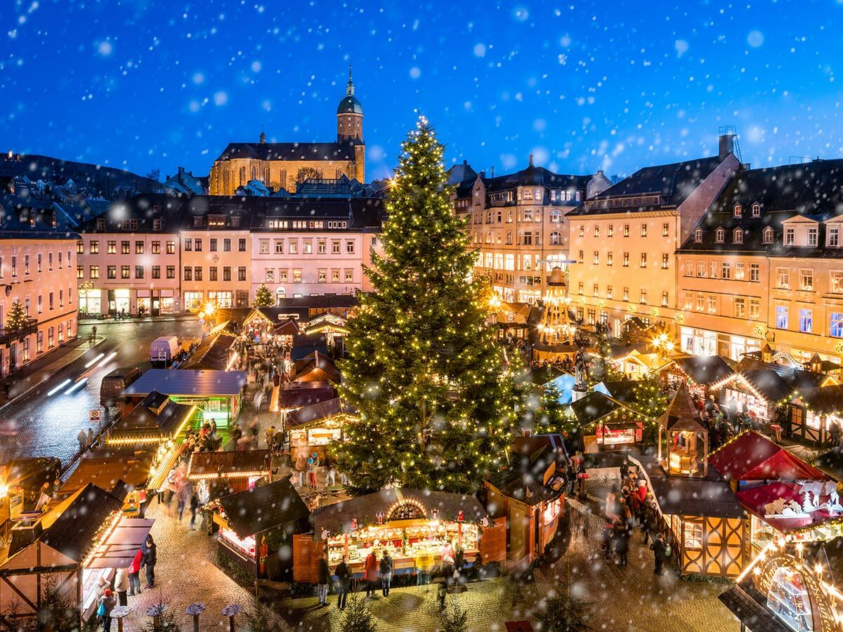 Festlicher Weihnachtsmarkt im Schnee, Abenddämmerung. Beleuchtete Stände, Menschen auf historischem Stadtplatz mit Kirche.