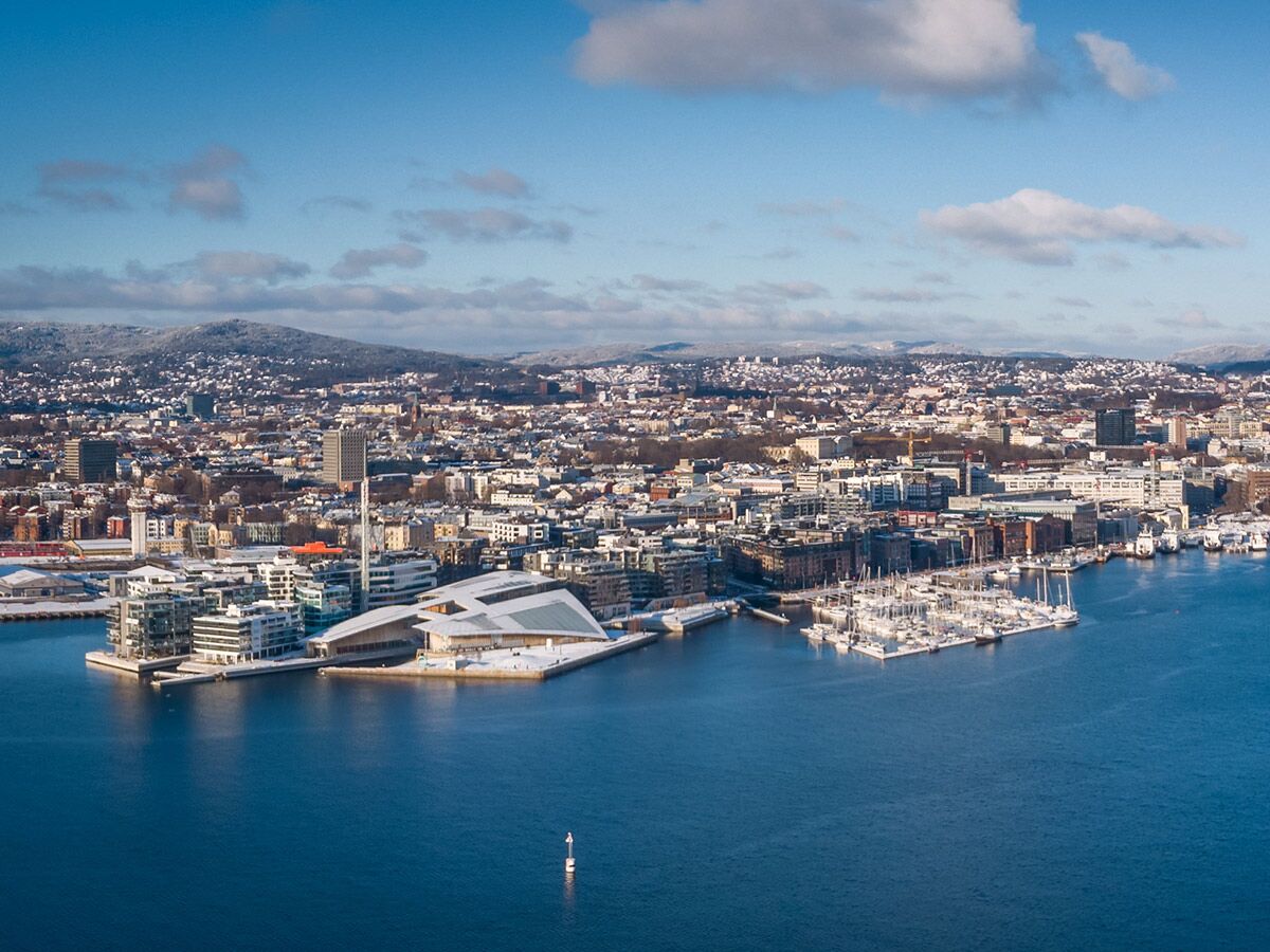 Luftbild von Oslo im Winter mit schneebedeckter Stadt, modernem Opernhaus, Yachthafen und Bergen im Hintergrund.