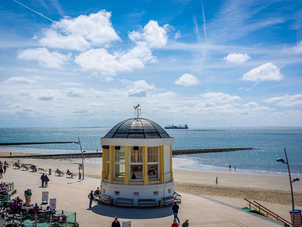 Blick auf belebte Strandpromenade mit gelb-weißem Rundgebäude, weitem Meer, Sandstrand, Menschen und Schiff unter blauem Himmel.