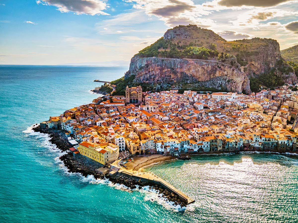 Luftaufnahme von Cefalù, Sizilien, mit bunten Häusern, Kathedrale, Felsen La Rocca und blauem Mittelmeer.