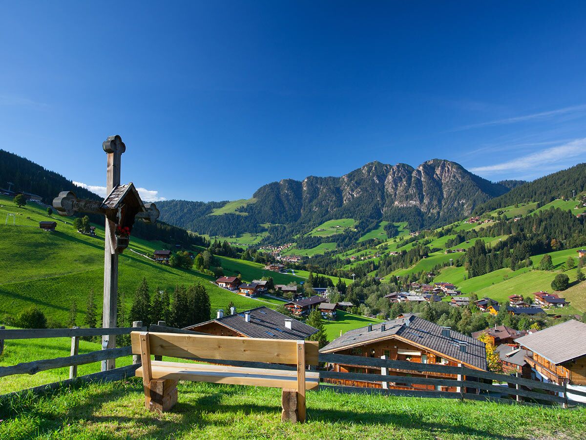 Idyllische Alpenlandschaft mit grünen Wiesen, einem Holzkreuz, einer Bank und einem Dorf im Tal vor Bergen unter blauem Himmel.
