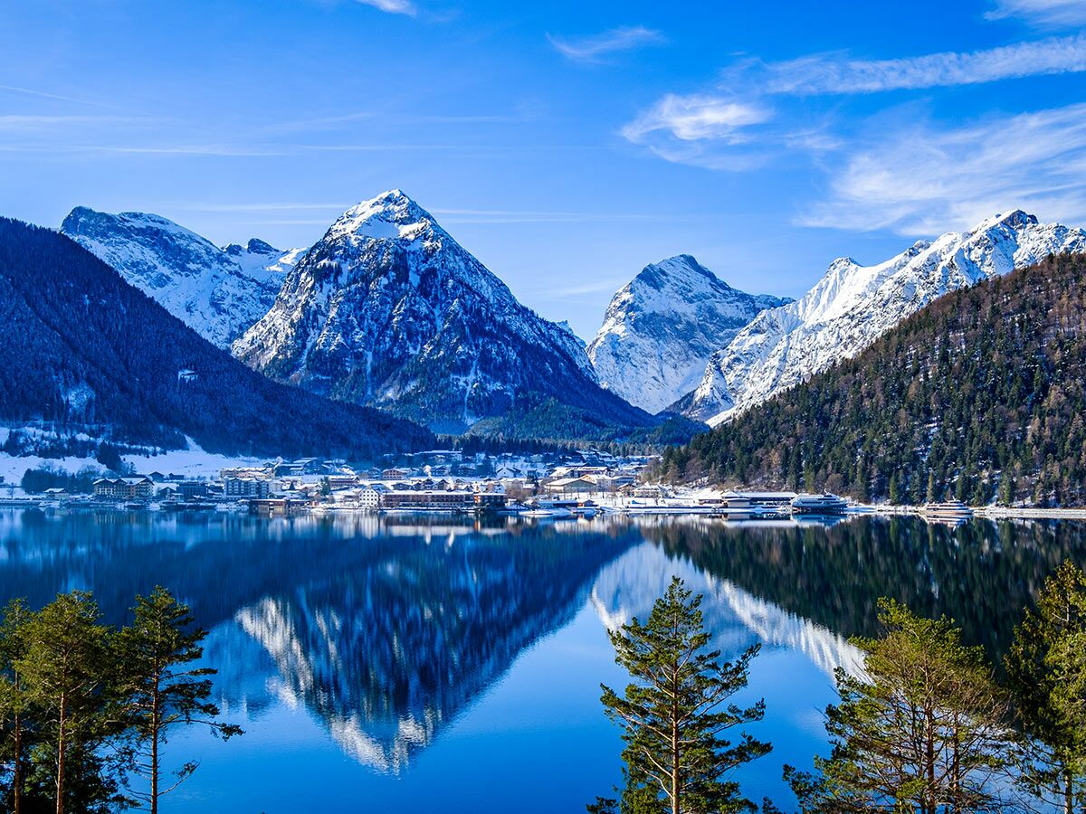 Idyllische Alpen-Winterlandschaft mit schneebedeckten Bergen, klarem See, Reflexion und charmantem Dorf am Ufer.