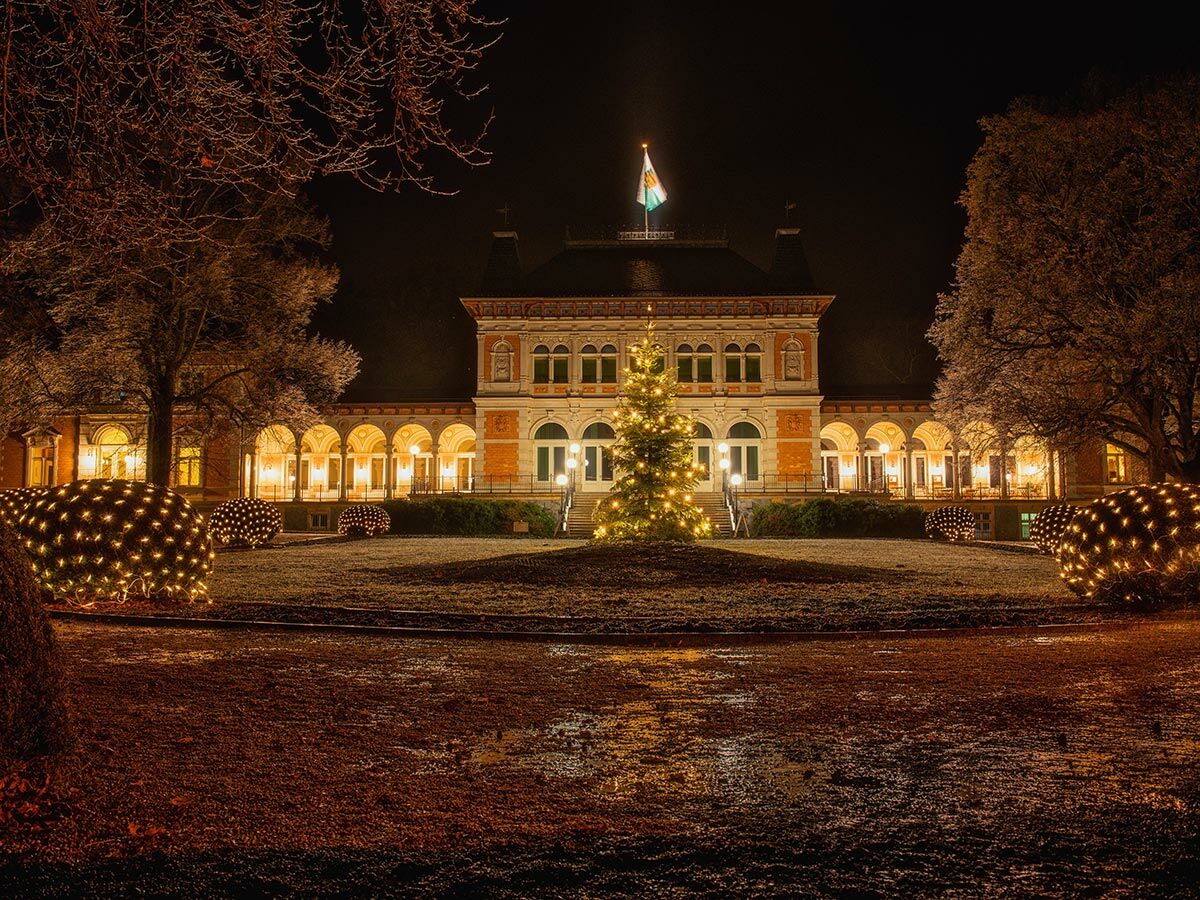 Historisches Schloss bei Nacht, festlich beleuchtet mit einem großen Weihnachtsbaum und Lichterketten im verschneiten Wintergarten.