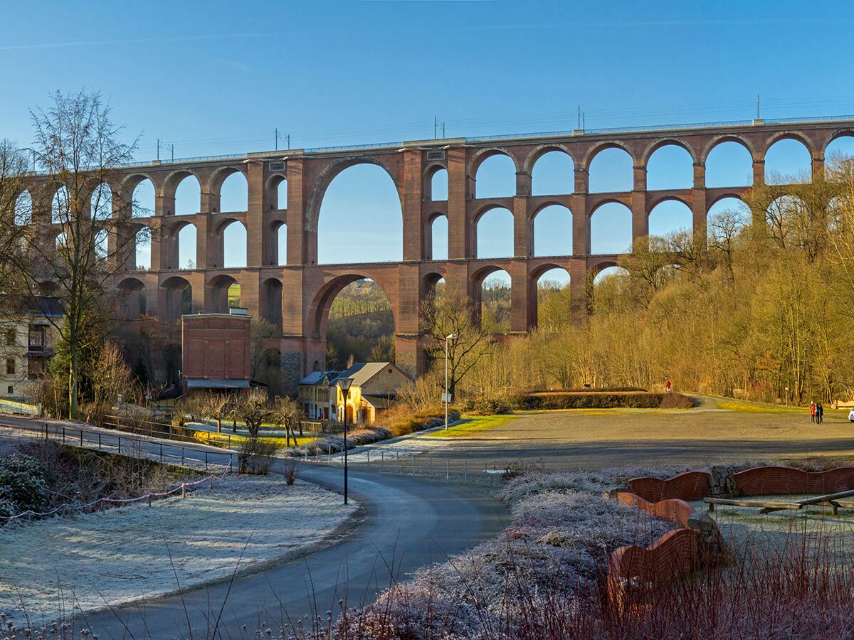 Imposantes dreistöckiges Ziegel-Viadukt mit vielen Bögen überspannt ein Tal mit Häusern und gefrorener Landschaft an einem sonnigen Wintertag.