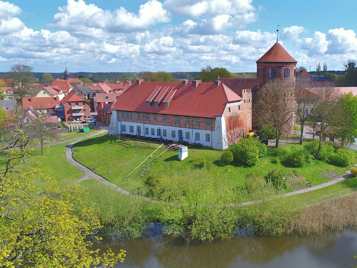 Luftaufnahme eines historischen Schlosses mit Turm, roten Dächern, See, grünen Wiesen und einem Dorf im Hintergrund bei strahlendem Himmel.