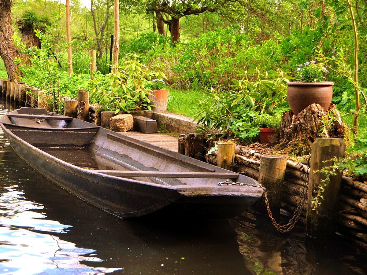 Dunkles Holzboot im grünen Kanal mit bewachsenem Ufer, Holzsteg und Pflanzen.