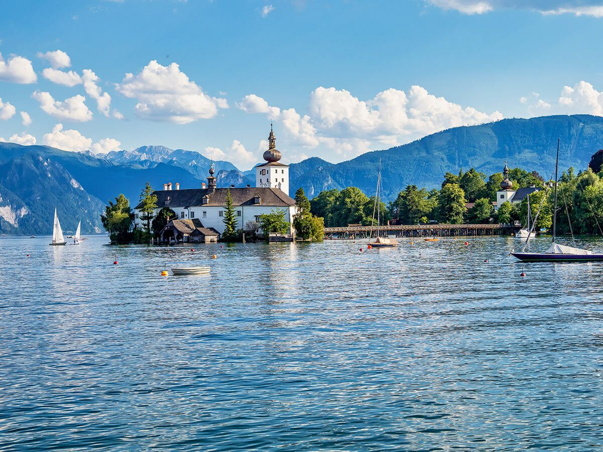 Malerisches Schloss Ort auf einer Insel im Traunsee bei Gmunden, Österreich, umgeben von Bergen und Segelbooten unter blauem Himmel.