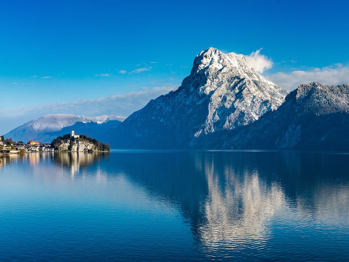 Panoramablick auf den winterlichen Traunsee mit dem idyllischen Schloss Ort auf einer Insel und dem schneebedeckten Traunstein im Hintergrund.