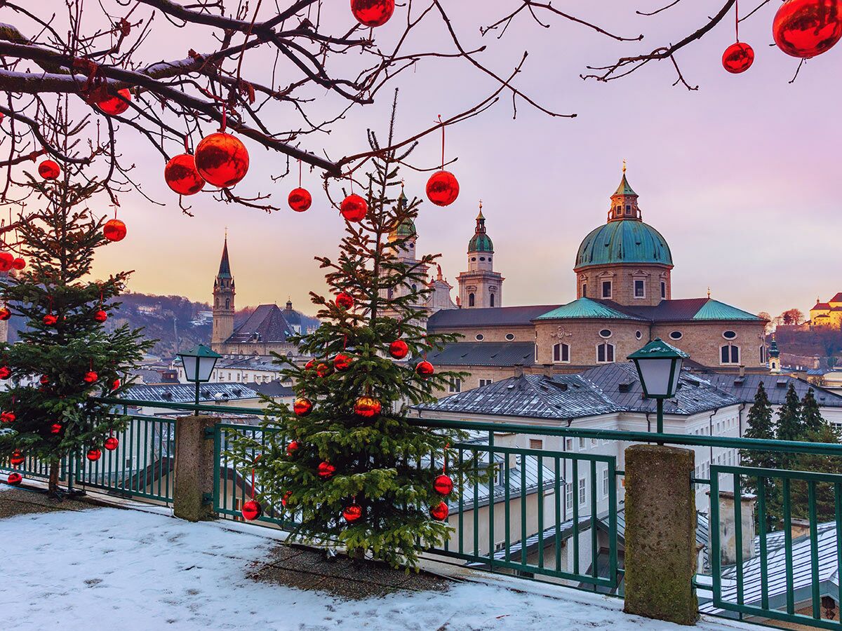 Winteransicht Salzburg: Christbäume und rote Kugeln vor Dom, Festung Hohensalzburg und schneebedeckter Stadt bei Sonnenuntergang.