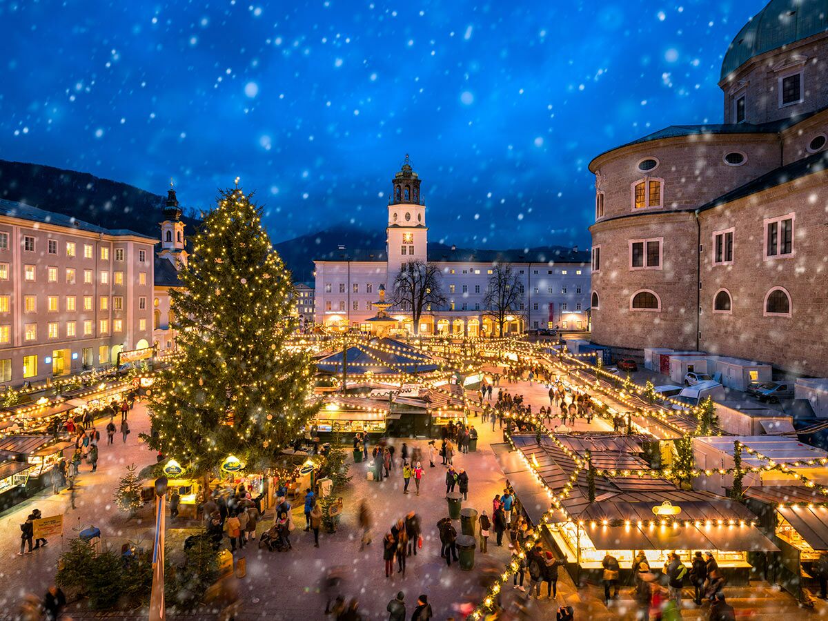 Salzburger Christkindlmarkt bei winterlicher Nacht mit Schneefall, geschmücktem Baum, beleuchteten Ständen und vielen Besuchern.