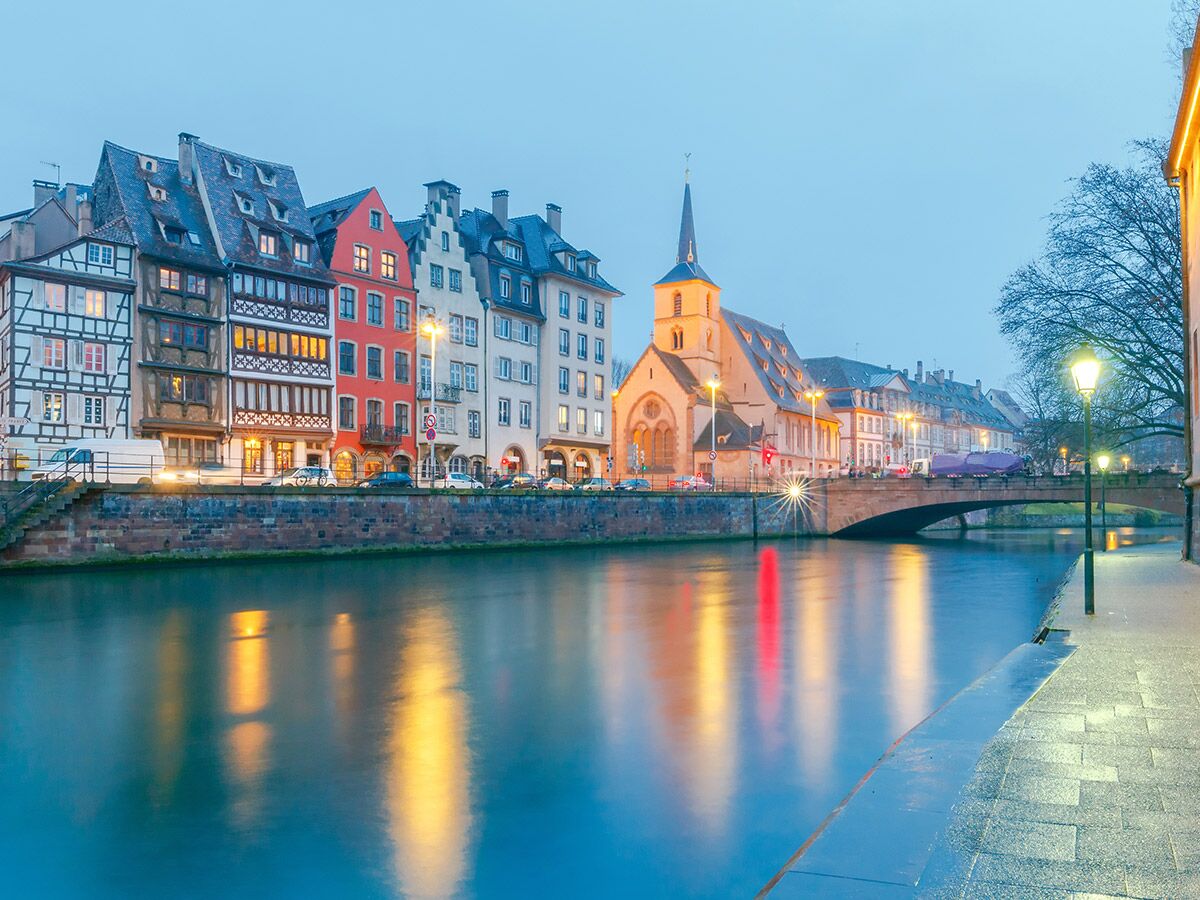 Romantische Abendansicht von Straßburg mit beleuchteten Fachwerkhäusern, Kirche und Brücke am Fluss Ill, mit warmen Lichtreflexionen.