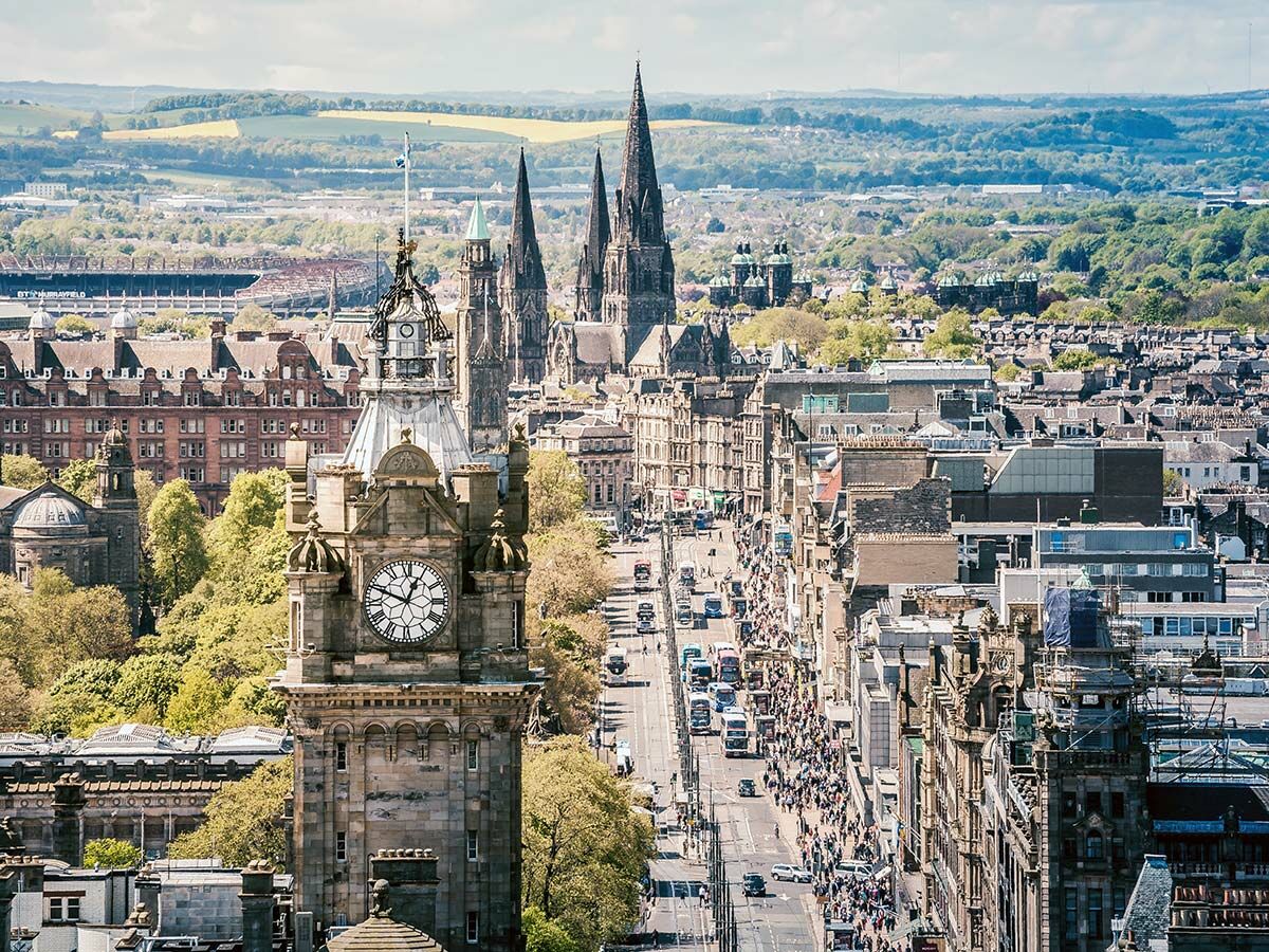 Weitläufige Stadtansicht von Edinburgh mit dem Balmoral Clock Tower, der belebten Princes Street, Menschen und Verkehr unter blauem Himmel.
