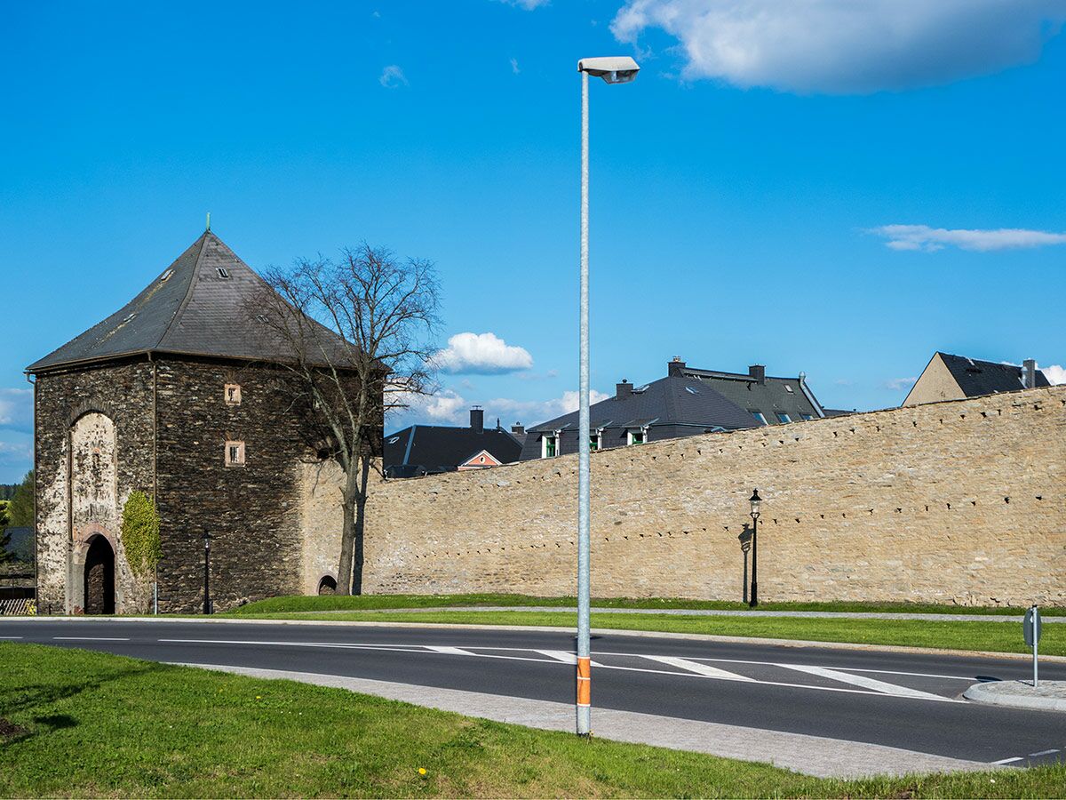 Alter Steinturm mit spitzem Dach und mittelalterlicher Stadtmauer neben einer Straße und Laterne unter blauem Himmel.