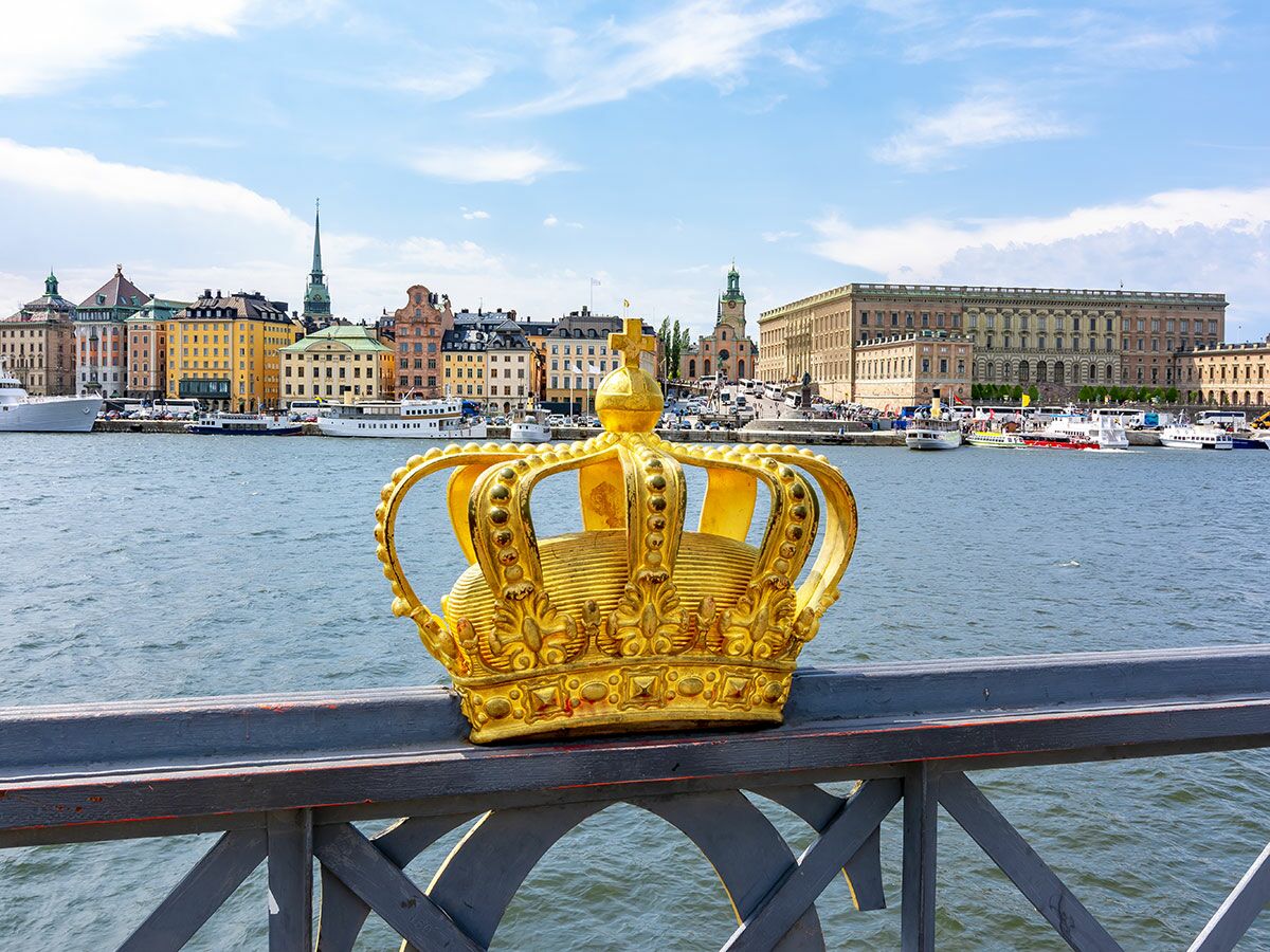 Goldene Krone auf Brückengeländer mit Blick auf die historische Stockholmer Stadtskyline, den Königlichen Palast und Boote auf dem Wasser.
