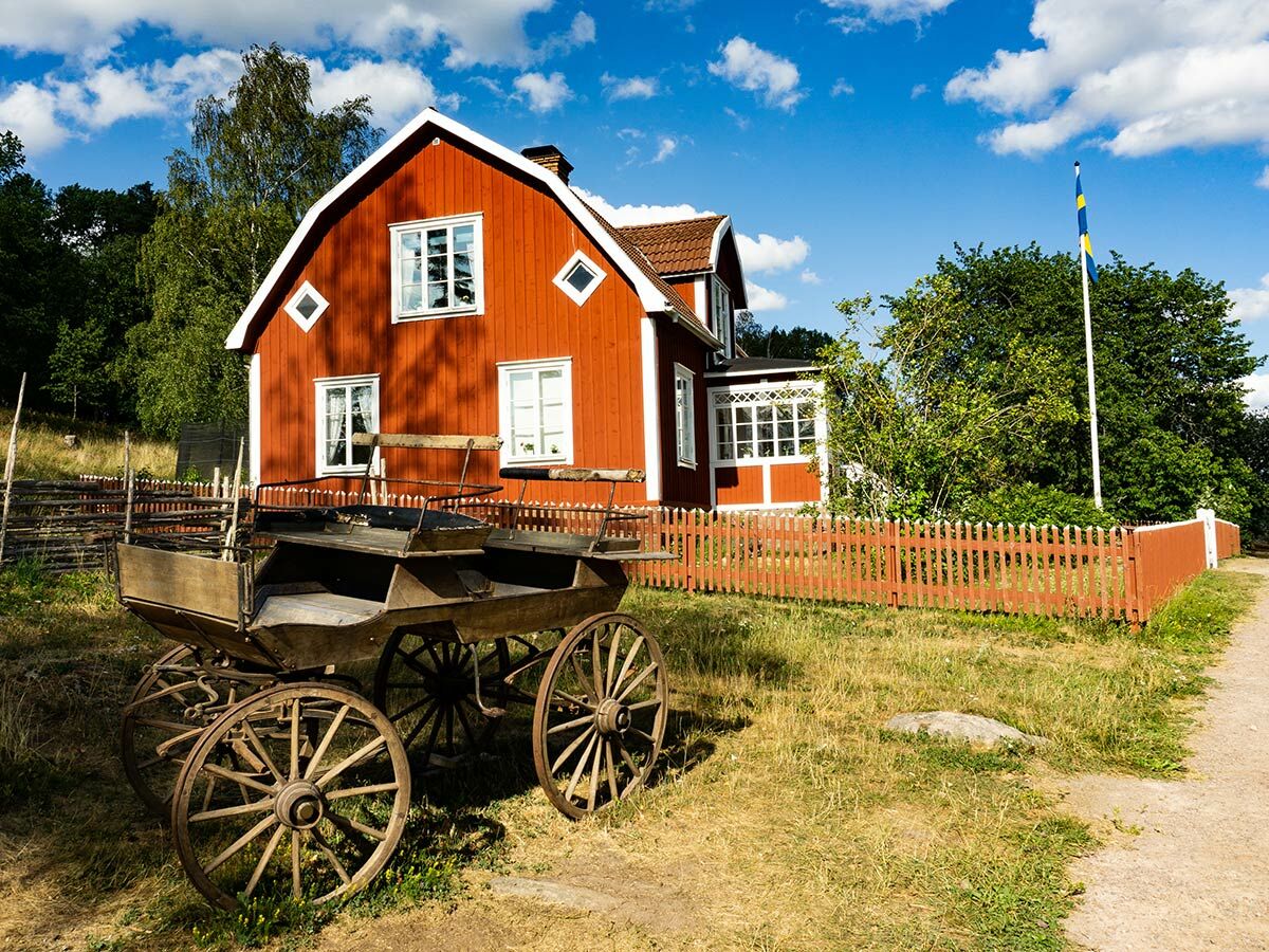 Traditionelles rotes Schwedenhaus mit weißen Fensterrahmen, einem alten Holzwagen, Holzzaun und einer wehenden schwedischen Flagge unter blauem Himmel.