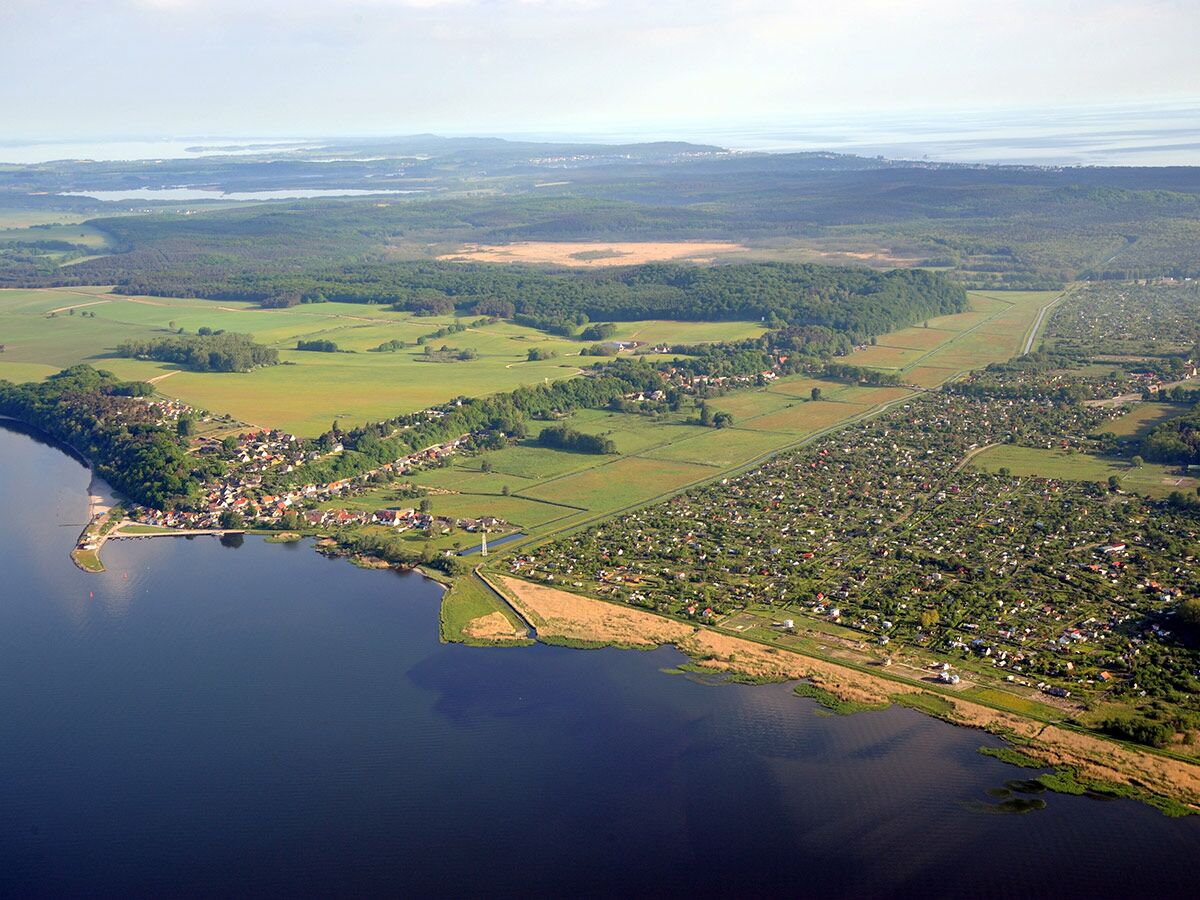 Luftaufnahme einer malerischen Küstenlandschaft mit einem Dorf, Feldern, Wäldern und weitläufigen Kleingartenanlagen am Wasser.