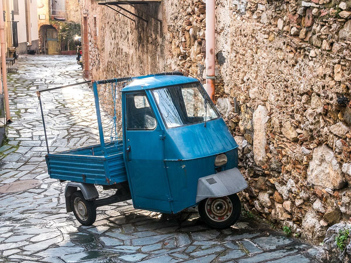 Ein kleiner, blauer Piaggio Ape Lieferwagen steht auf einer nassen, gepflasterten Gasse zwischen alten Steinmauern in Südeuropa.