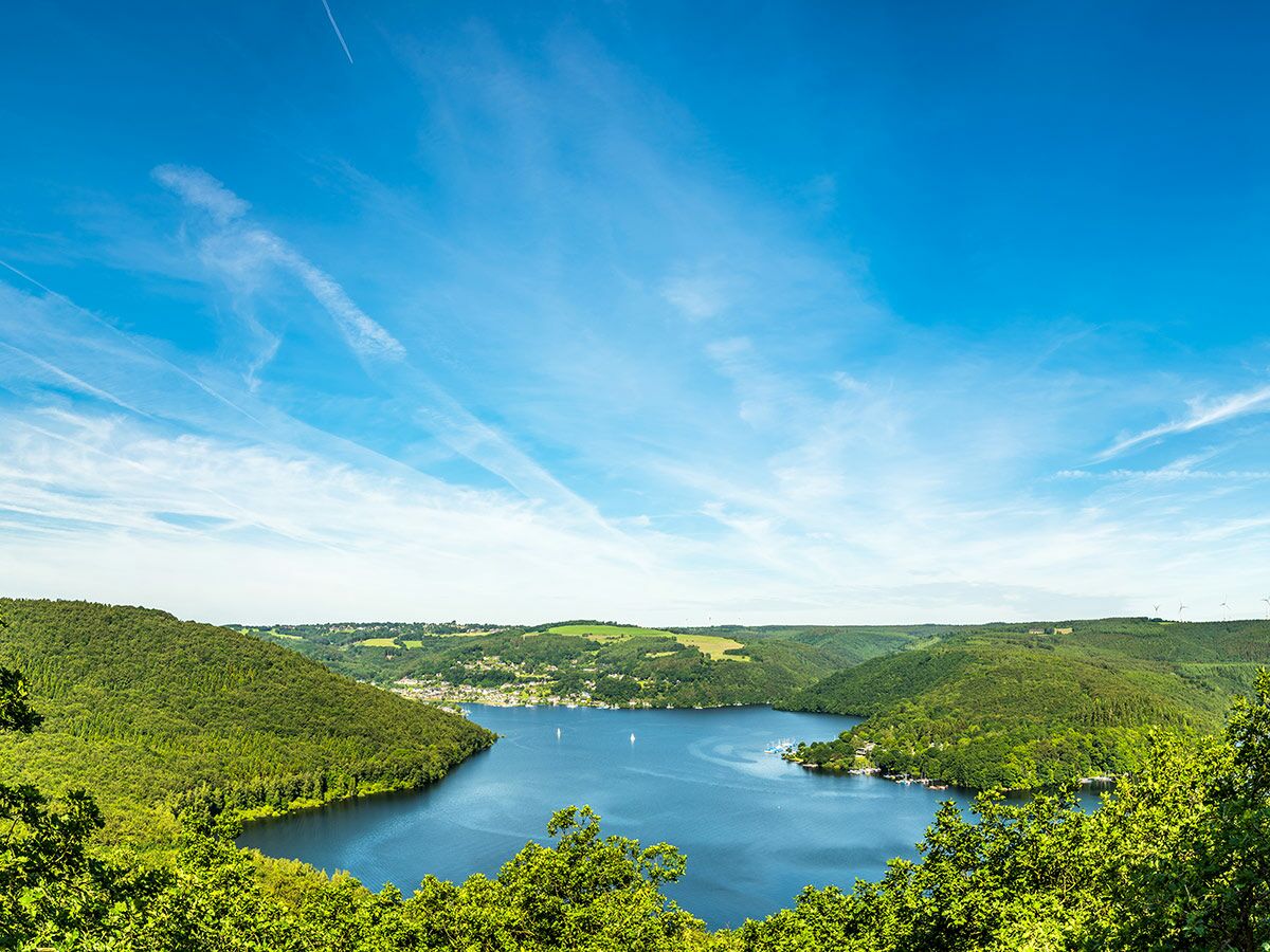 Weiter Blick auf ruhigen blauen See, umgeben von grünen bewaldeten Hügeln und einer Stadt unter klarem Himmel.