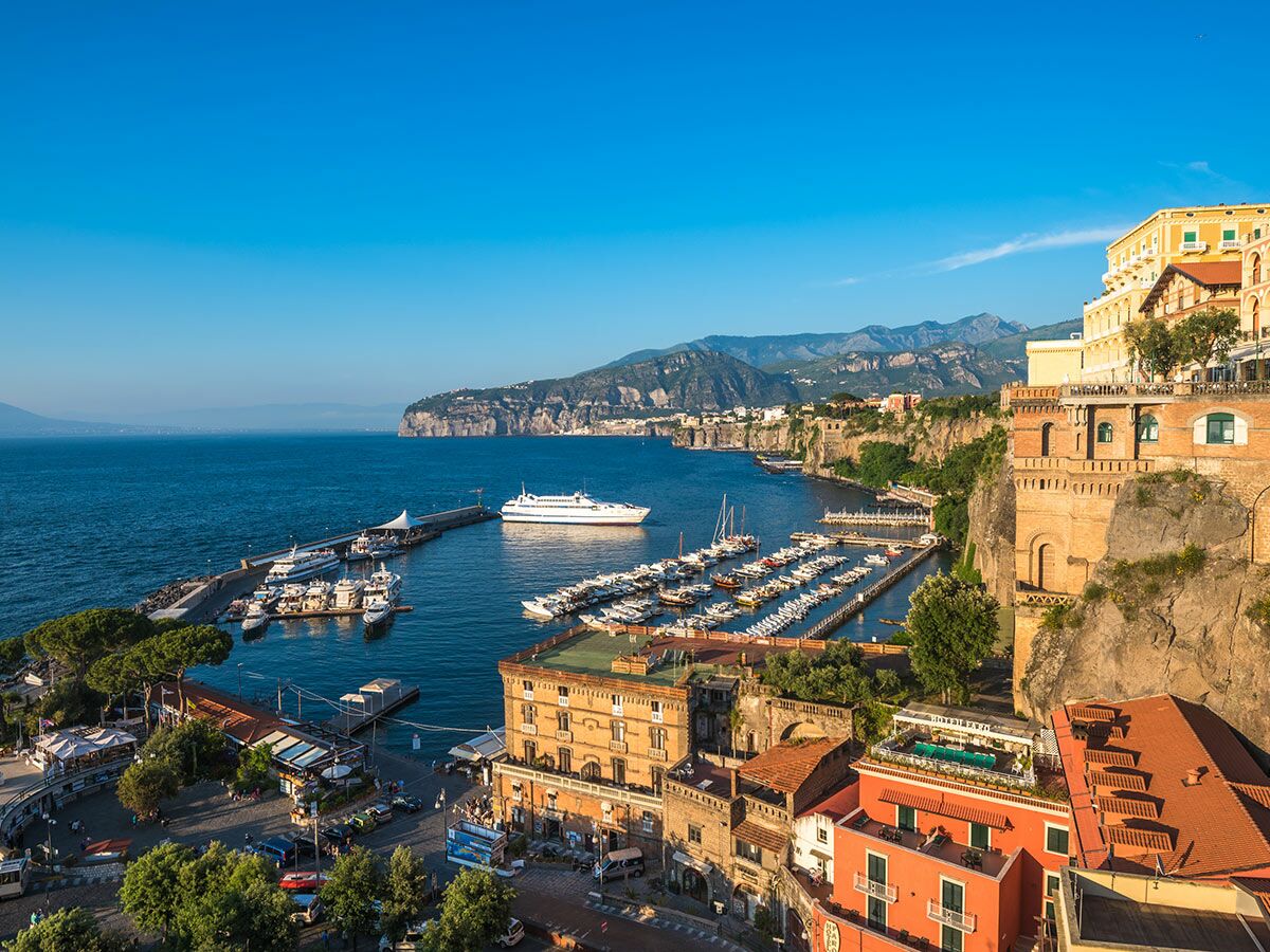 Panoramablick auf die Küstenstadt Sorrento in Italien mit Yachthafen, Fähre, bunten Klippenhäusern und blauem Mittelmeer.
